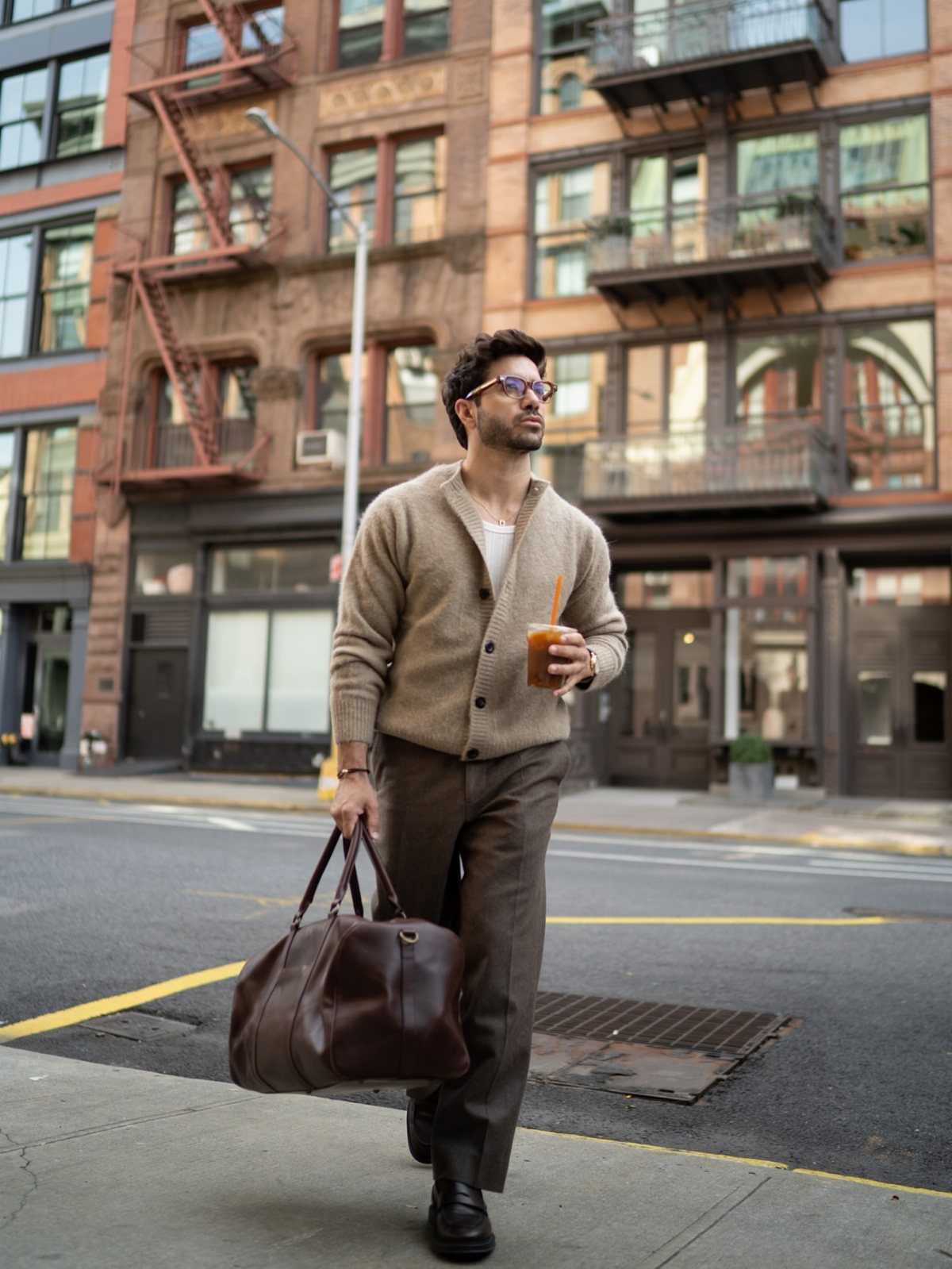 Man in a beige cardigan and brown trousers walking through a city street with a leather bag.
