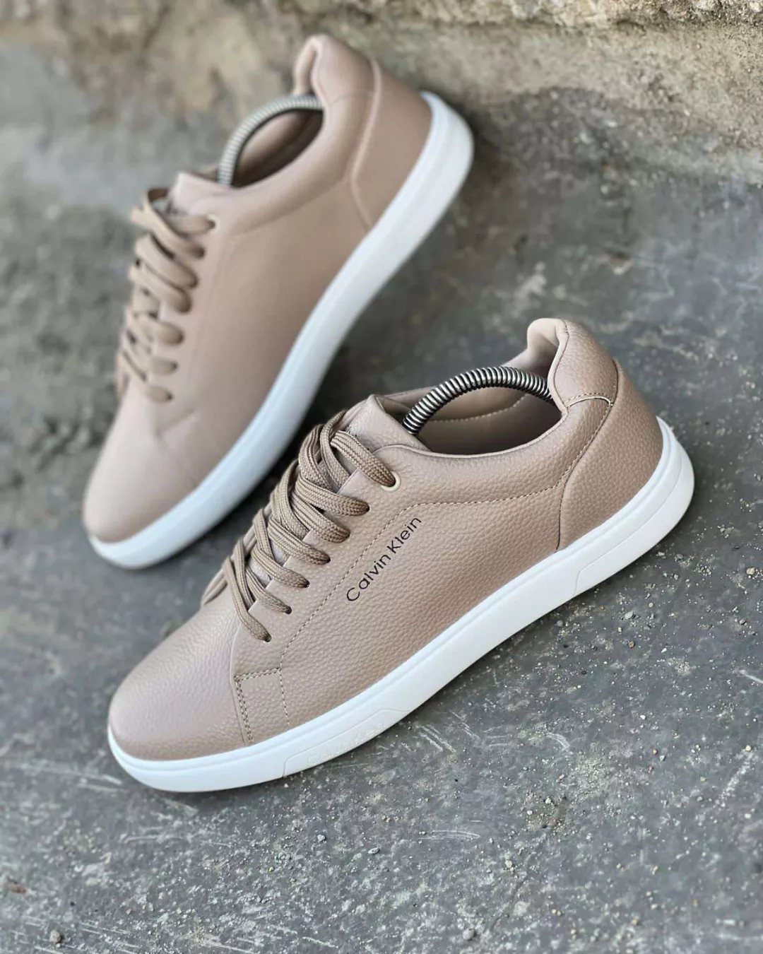 Beige Calvin Klein leather sneakers with white soles resting on a grey stone surface.