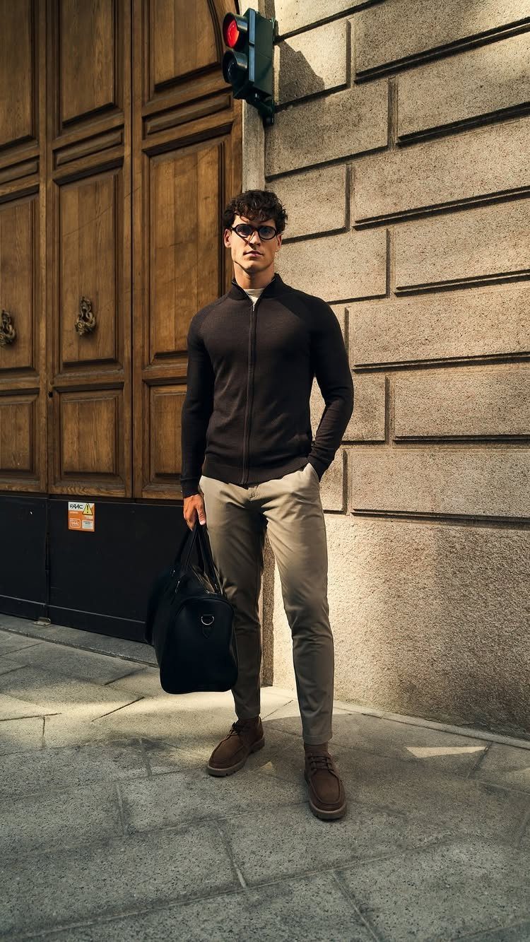 Man in a black zip-up sweater and grey chinos standing by a stone wall.