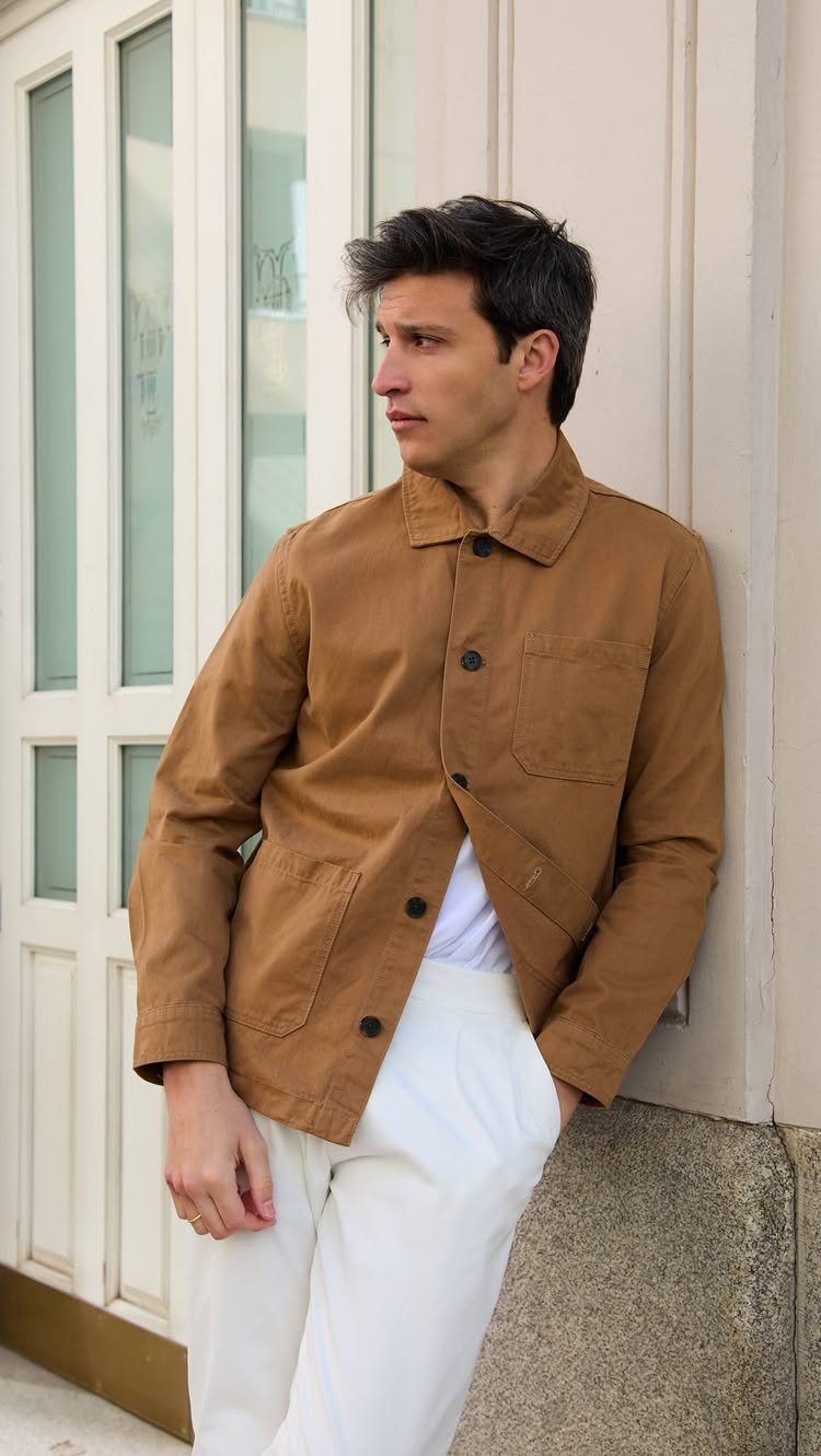 Man in a brown jacket and white trousers leaning against a white wall.