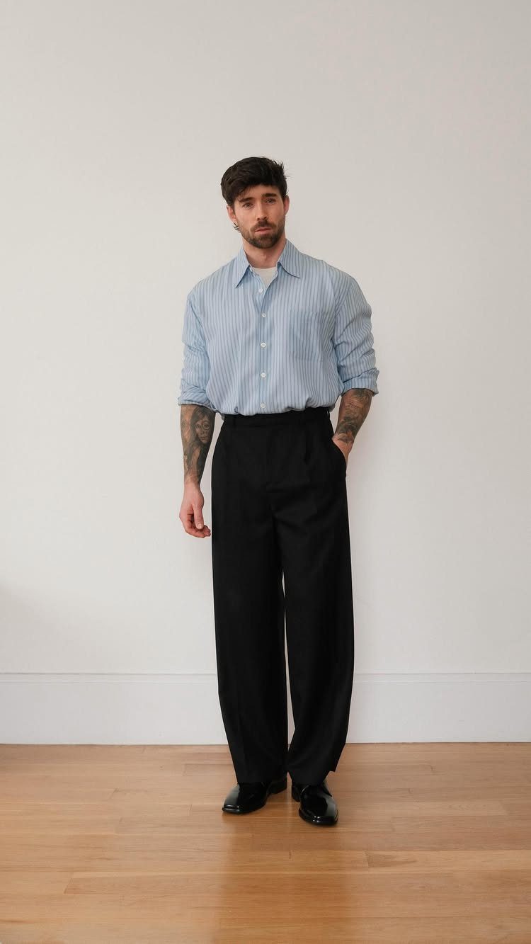 A man wearing a striped shirt, wide black trousers, and black leather shoes standing against a white wall.