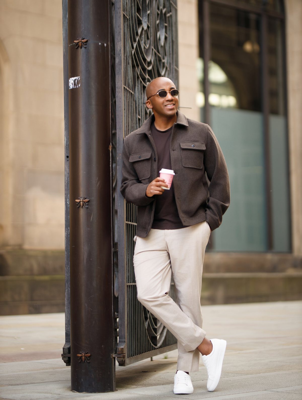 A man in a brown wool shacket and beige trousers leans against a pillar holding coffee.