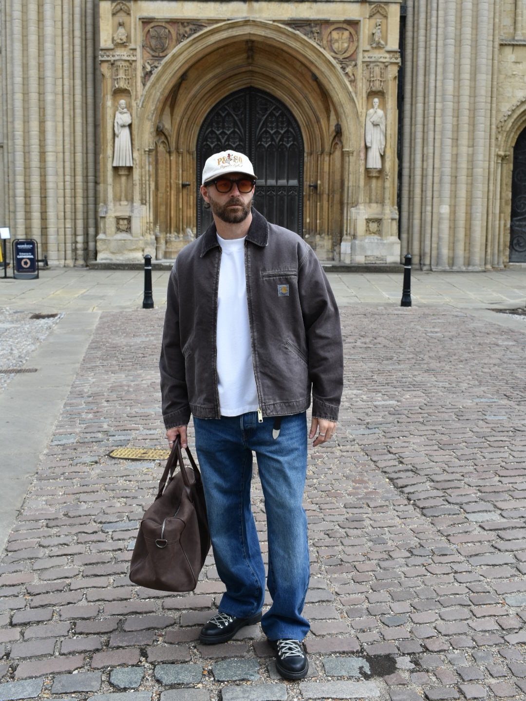 A man wearing a brown work jacket, blue jeans, and a white cap stands on a cobblestone street.