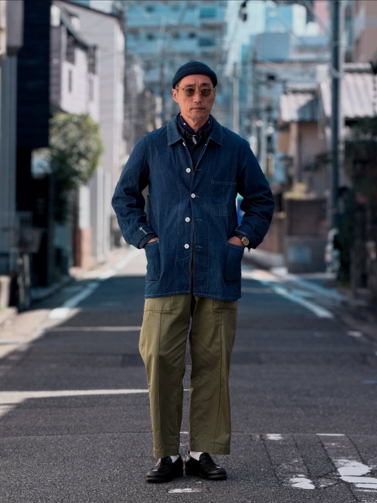 Man in denim jacket and olive green trousers standing on a narrow urban street.
