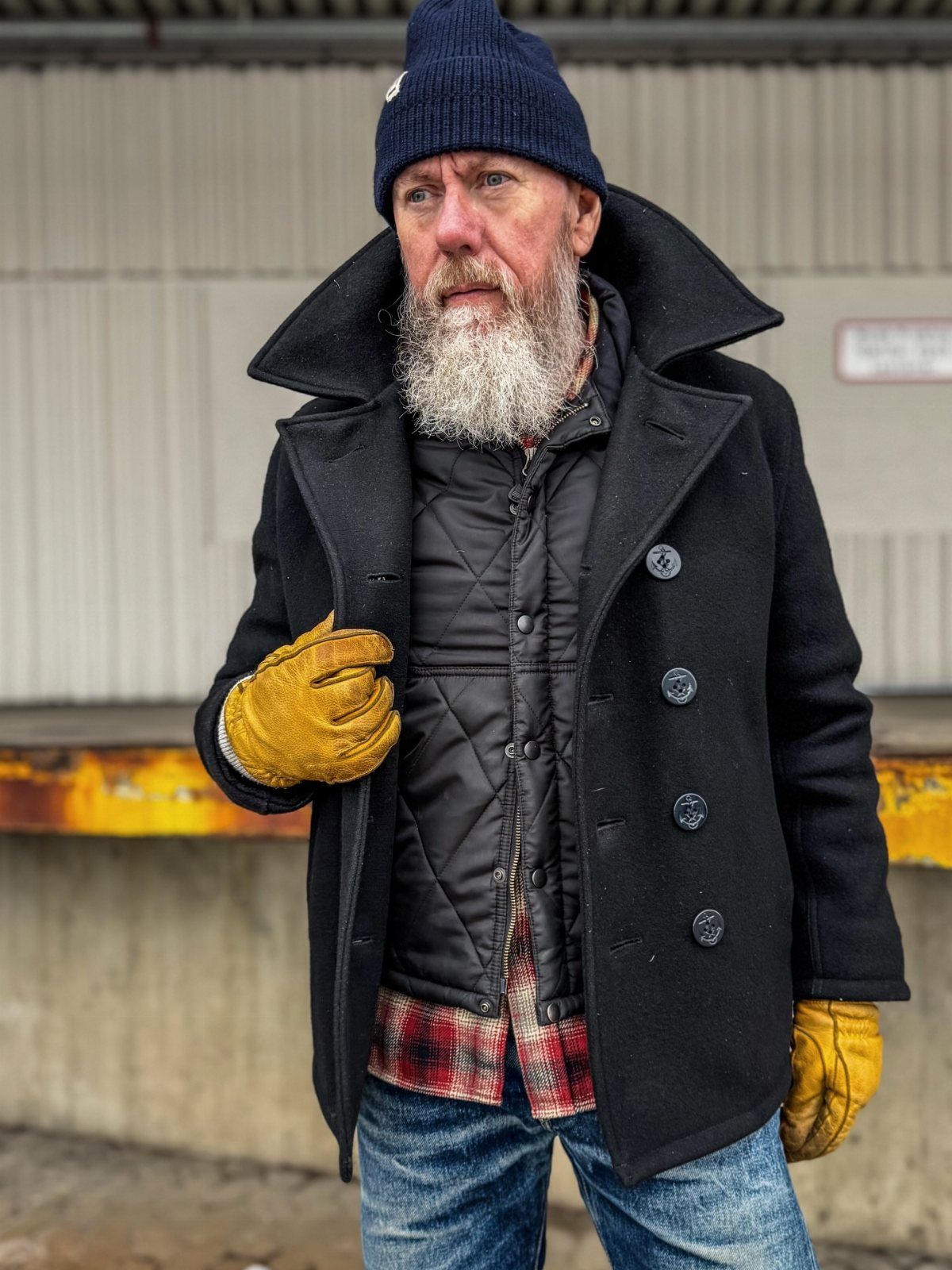 Bearded man in winter workwear including a black pea coat, quilted vest, and plaid flannel shirt.