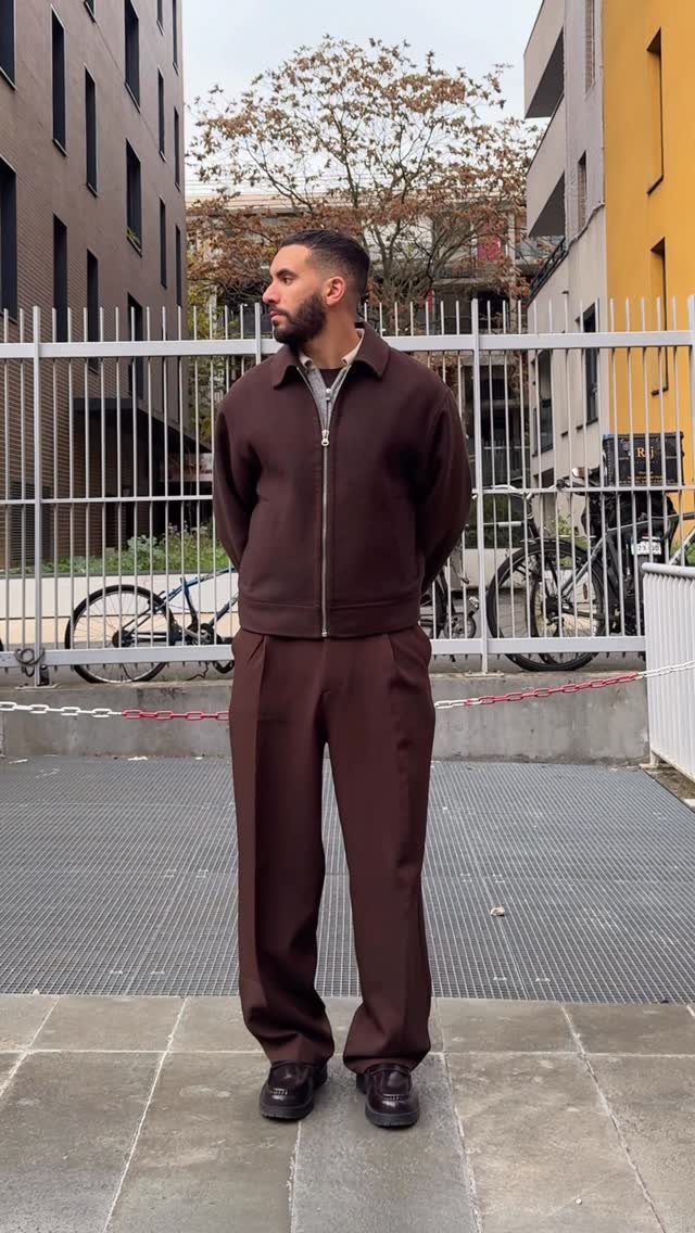 A man posing in a matching dark brown jacket and trousers set outdoors.