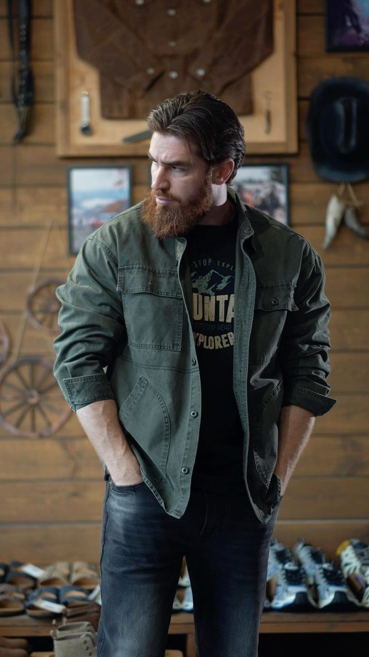 A bearded man wearing an olive green utility shirt and dark jeans standing in a rustic room.