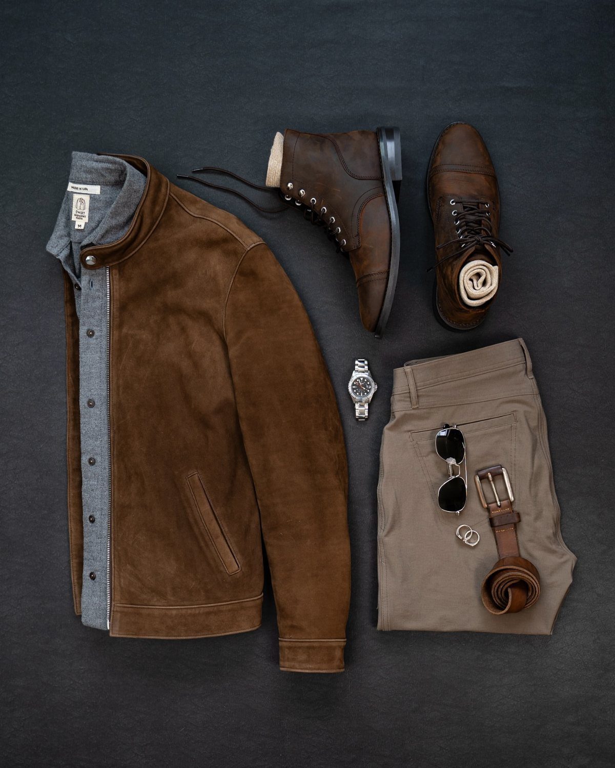 Flat lay of men's workwear including a brown suede jacket, leather boots, khaki pants, and accessories.