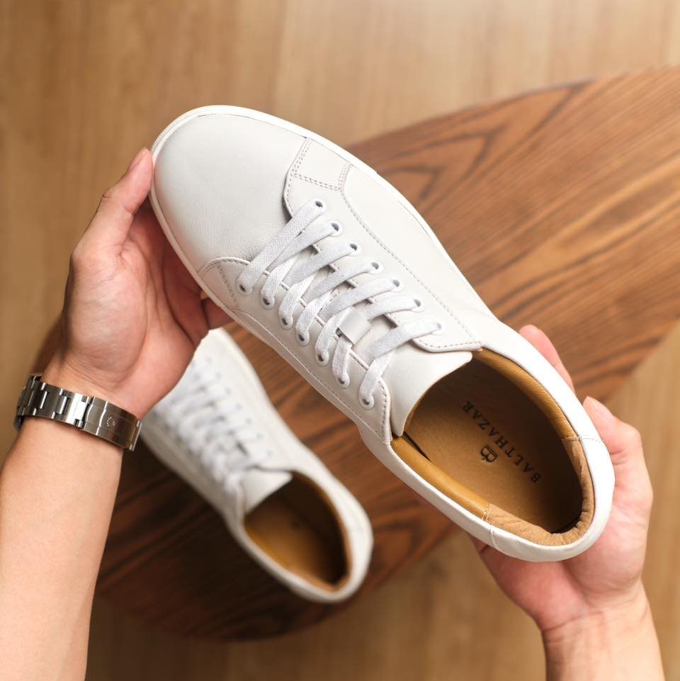 Hands holding a pair of minimalist white leather sneakers over a wooden table.