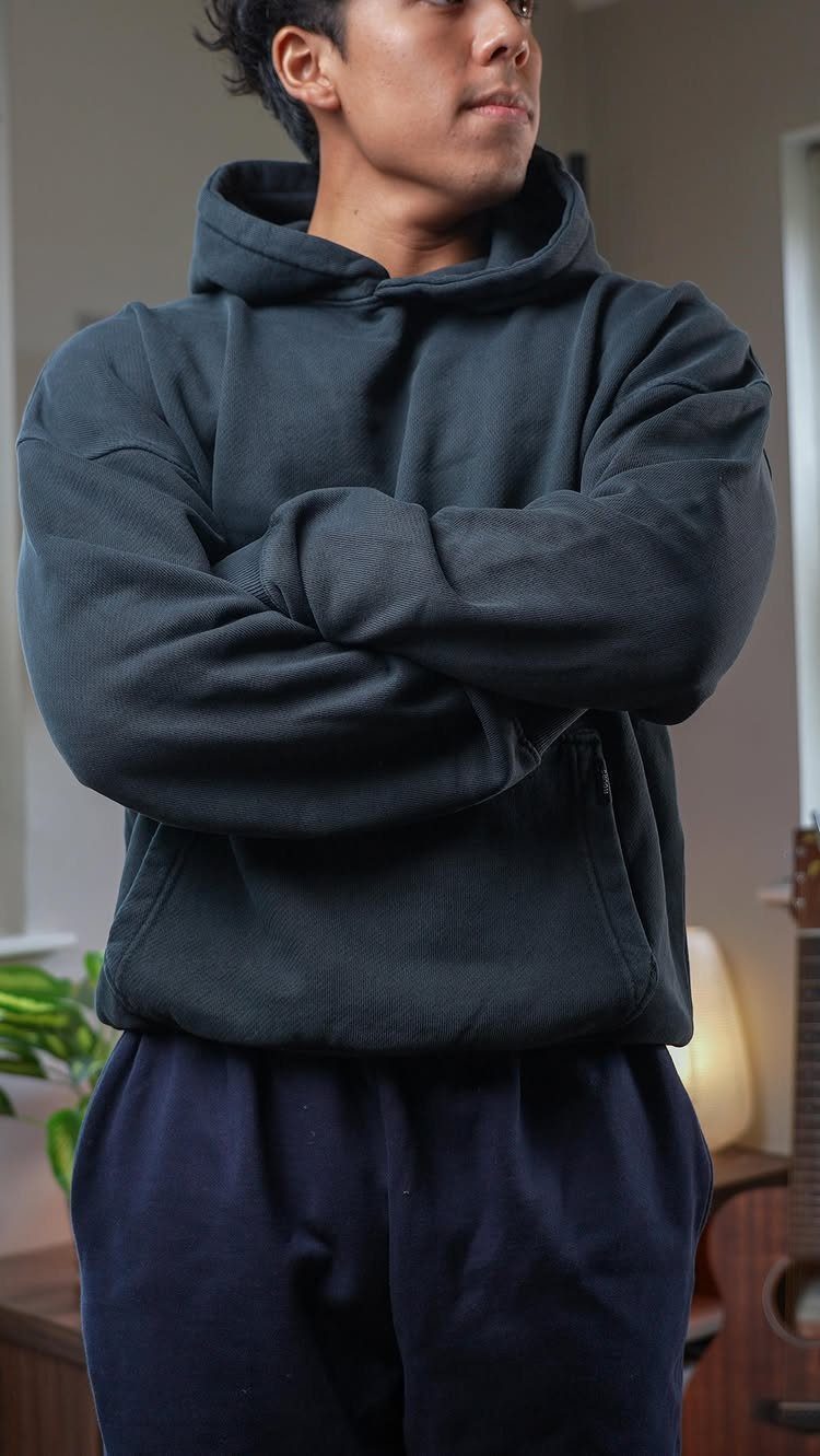 A man wearing a dark charcoal textured hoodie and navy blue sweatpants standing with arms crossed.