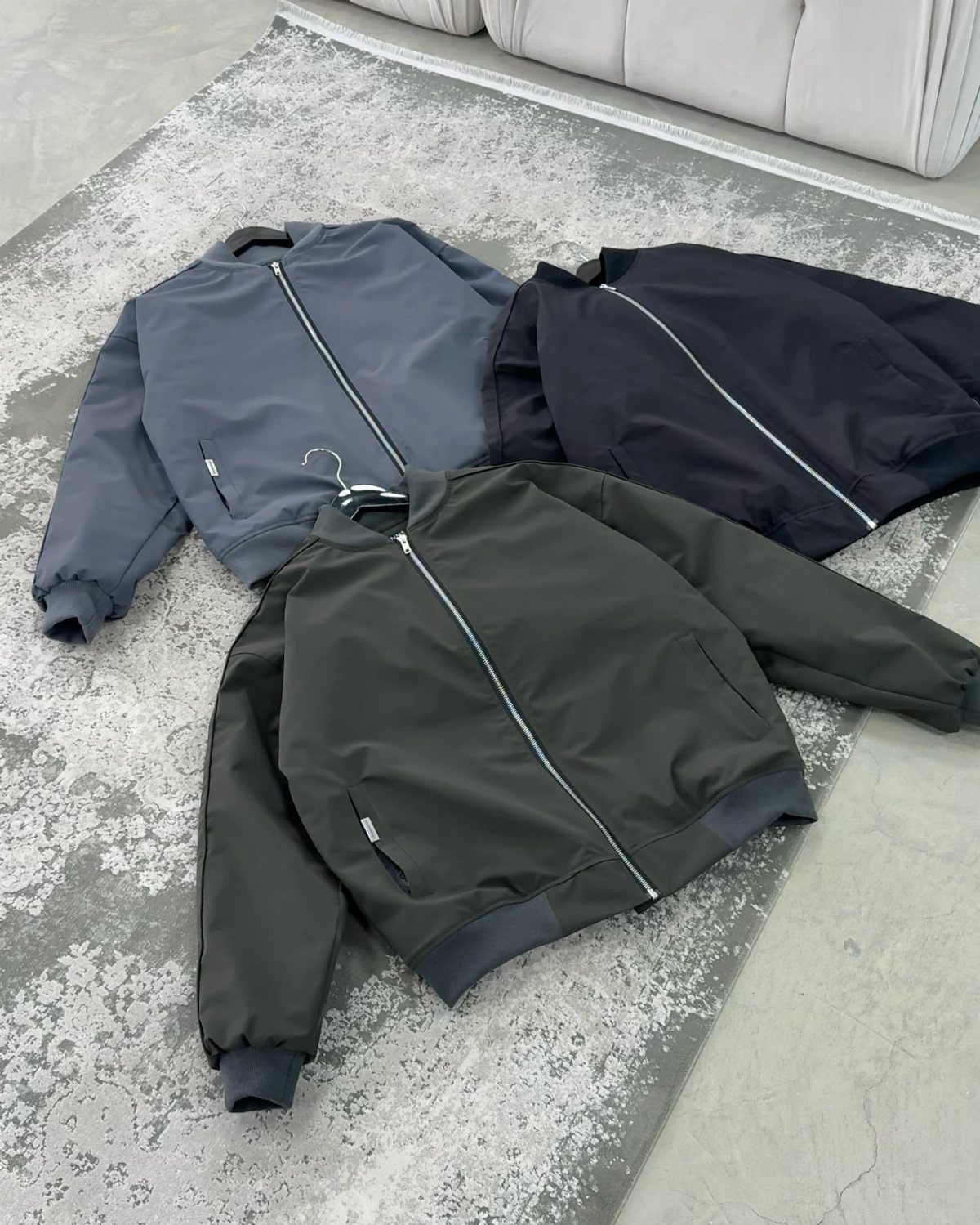 Three minimalist zip-up bomber jackets in blue, black, and olive green laid on a grey rug.