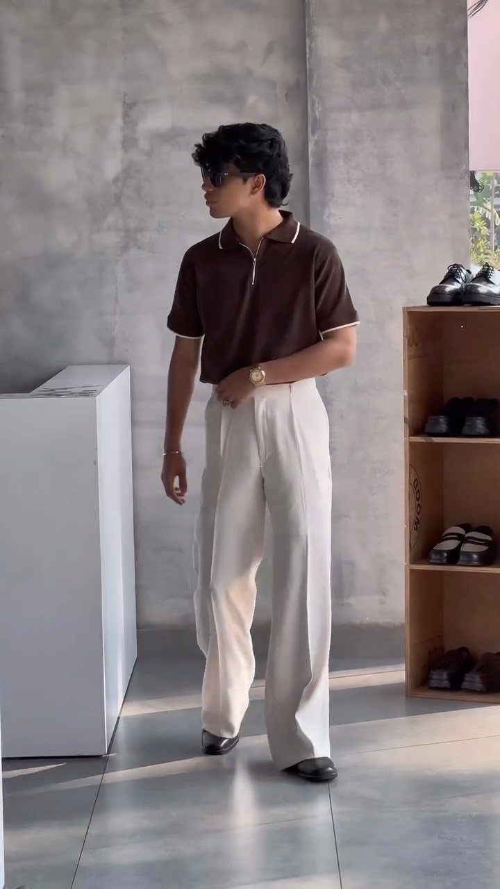 Man wearing a brown polo shirt and wide-leg cream trousers standing in a minimalist room.