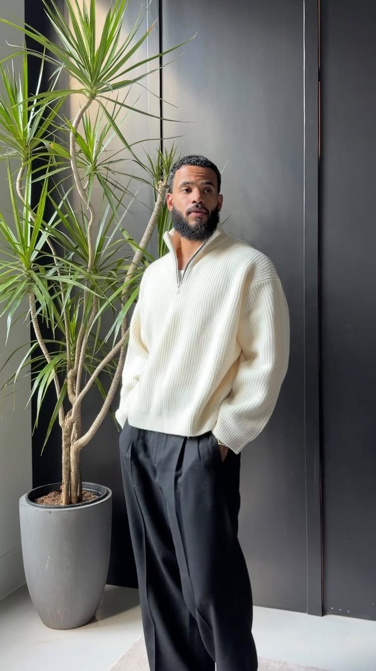 A man wearing a cream ribbed quarter-zip sweater and black trousers standing by a plant.