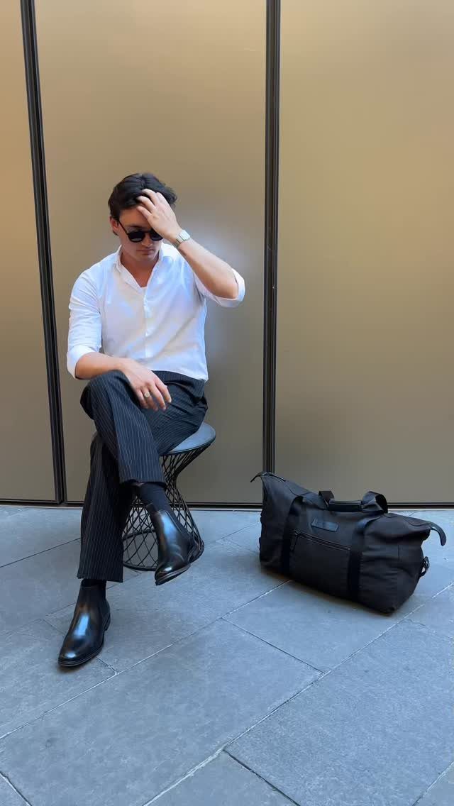 A man in a white shirt and black trousers sits on a stool next to a black bag.
