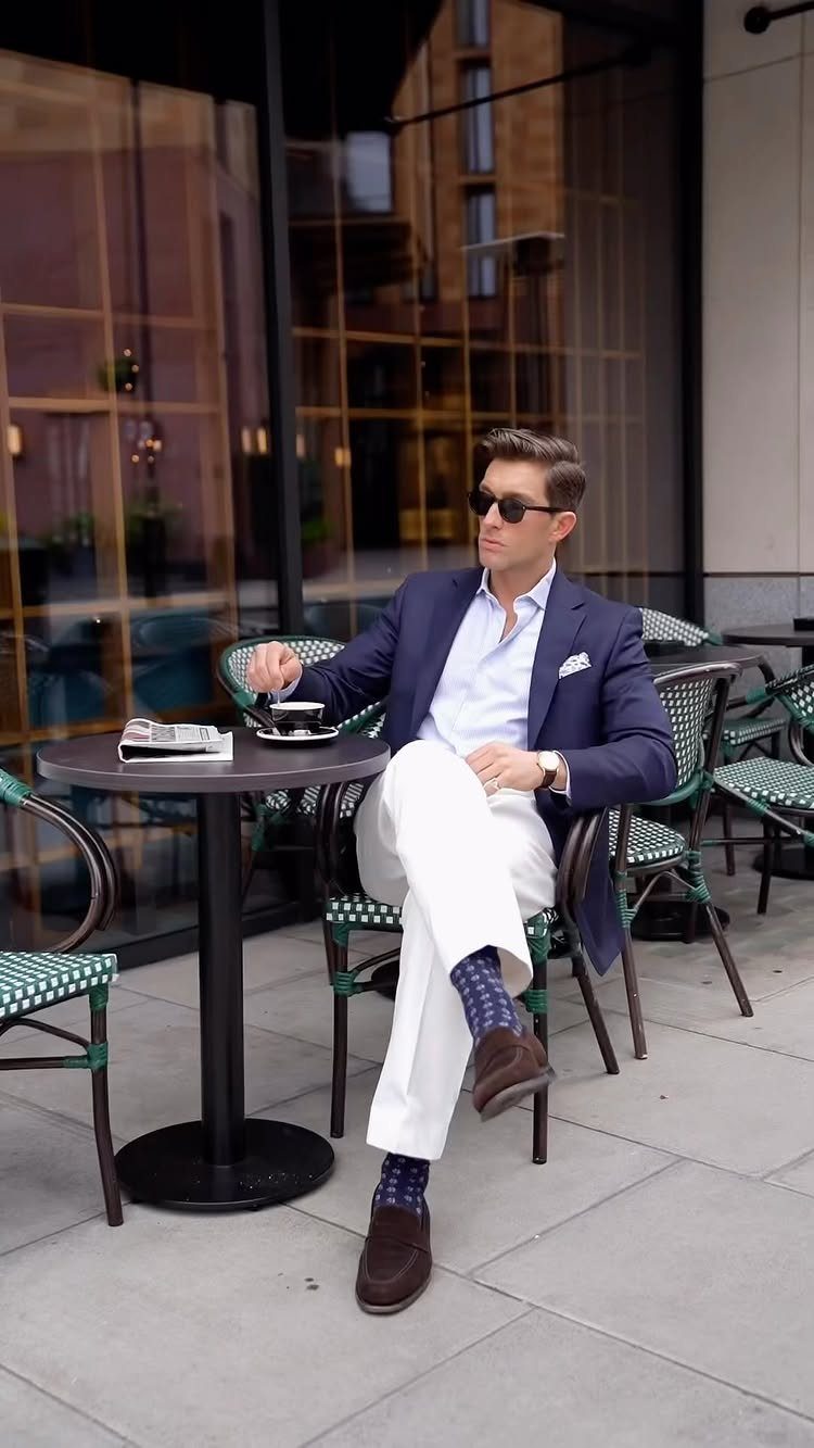 Man in navy blazer and white trousers sitting at an outdoor cafe table wearing sunglasses.