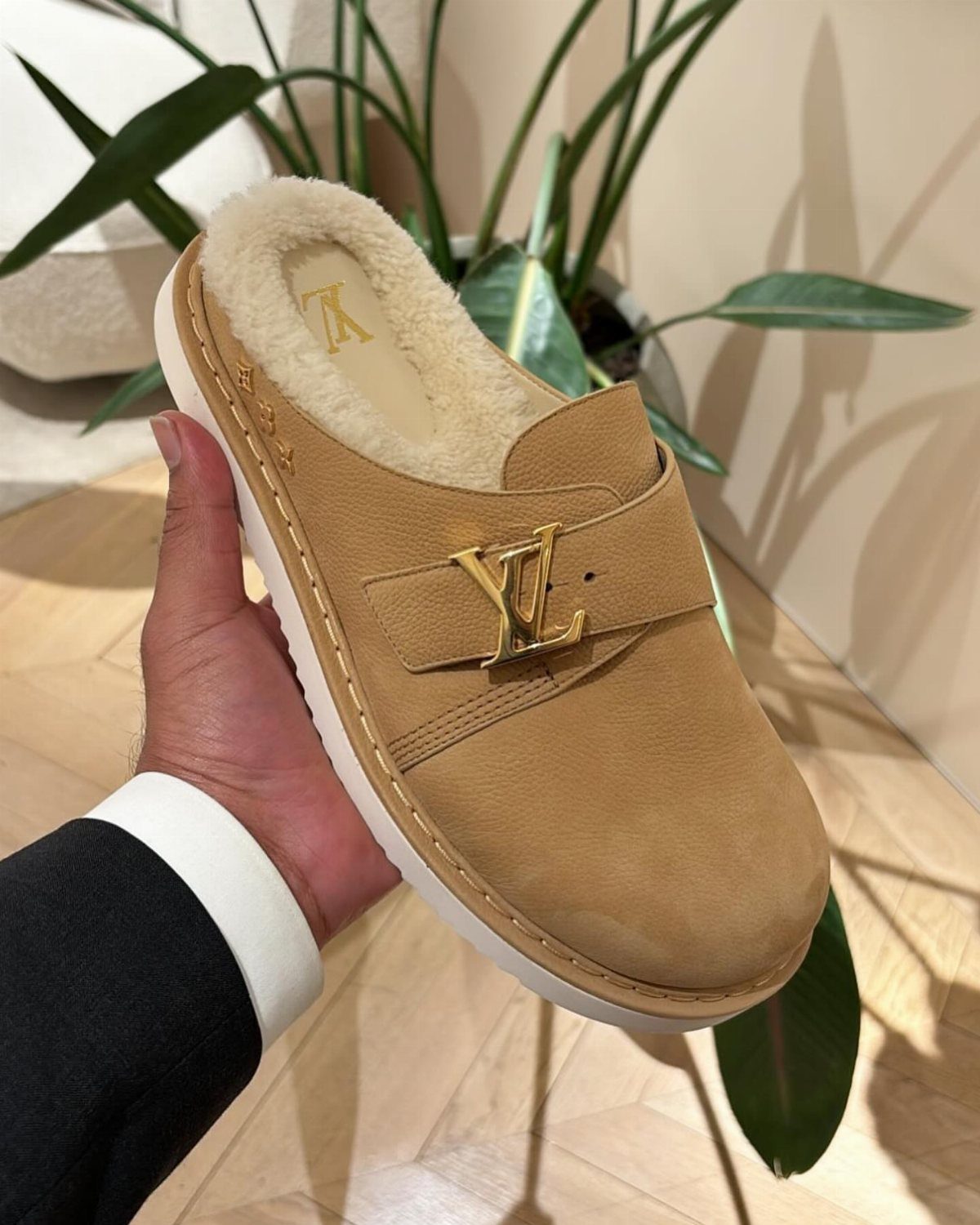 A hand holding a tan suede Louis Vuitton clog with cream shearling lining.