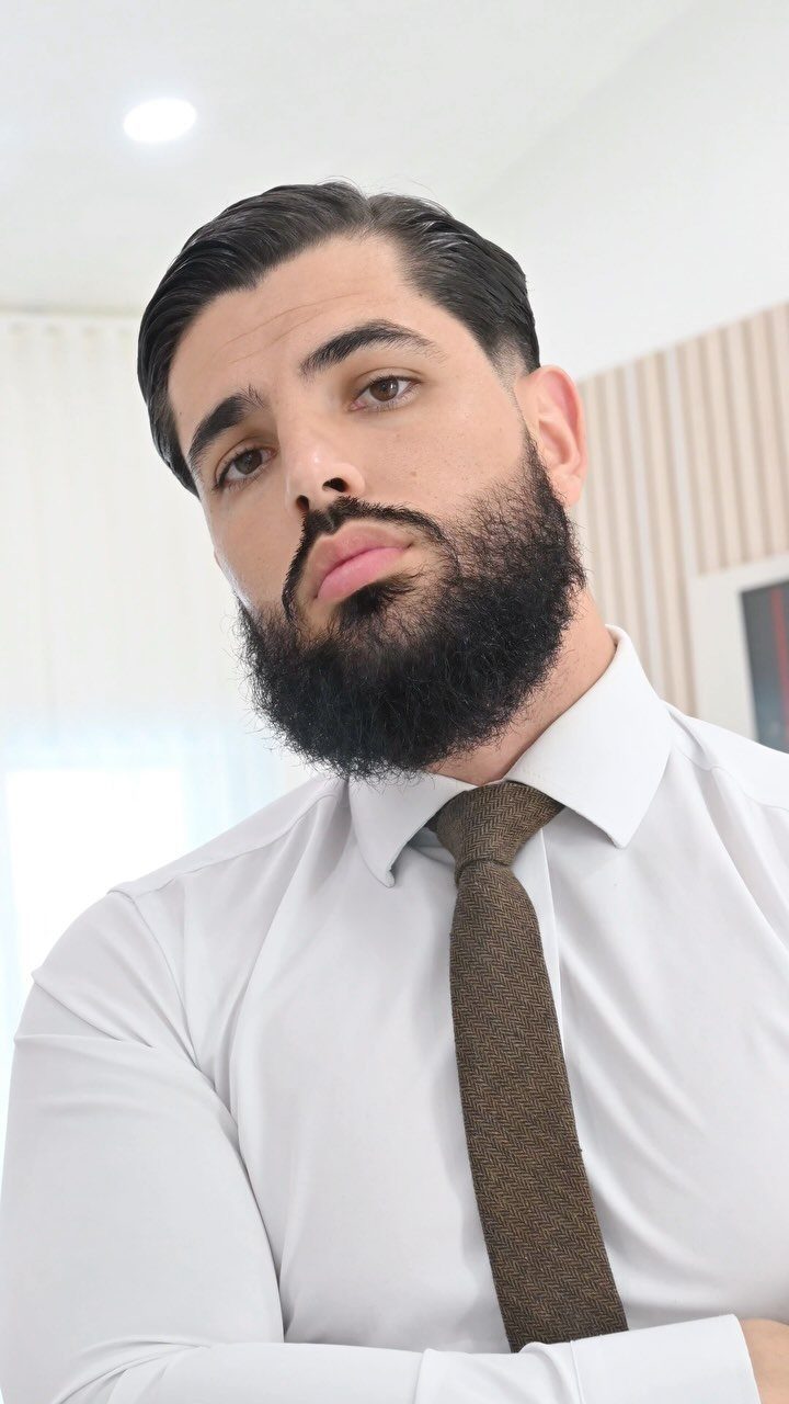 A man with slicked-back hair and a groomed beard wearing a white shirt and brown tie.