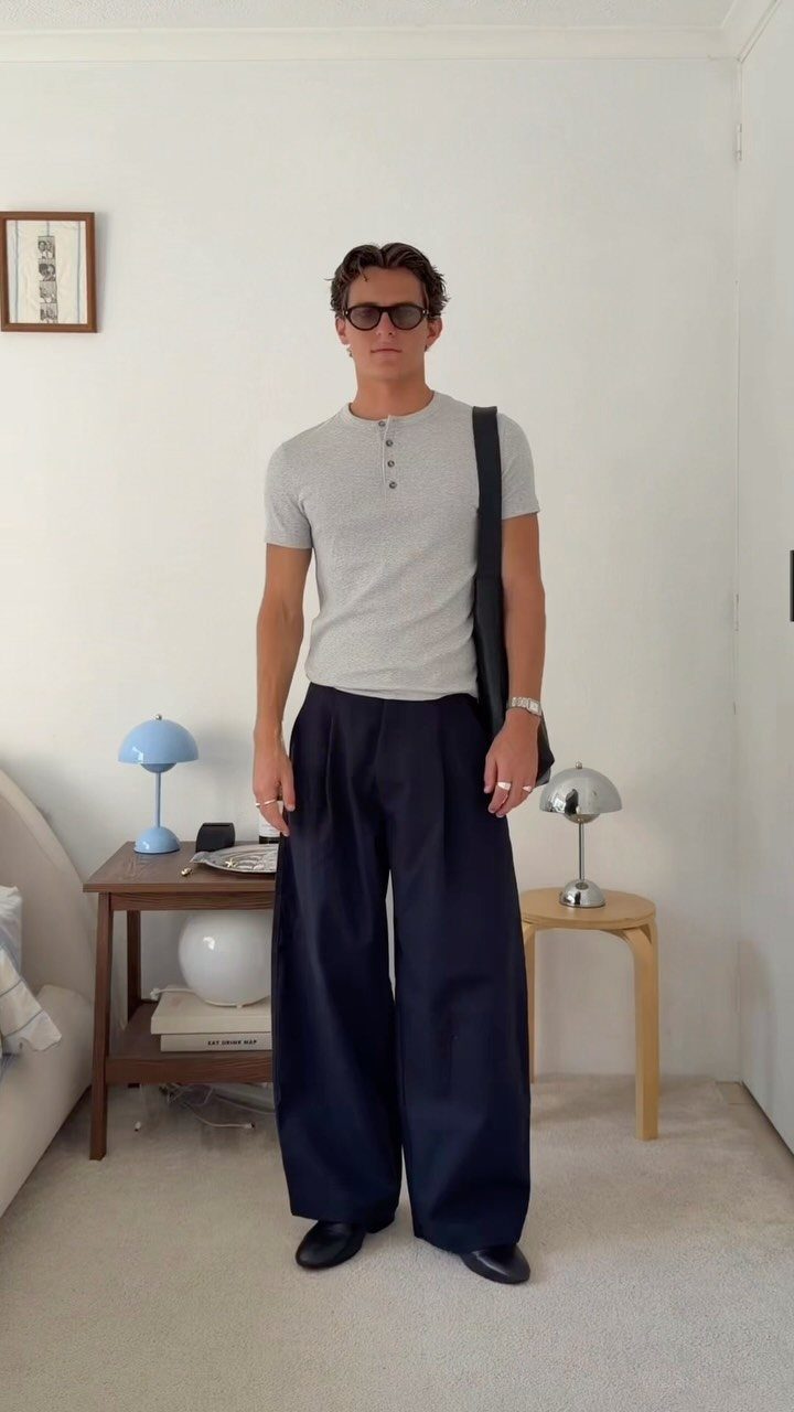 A man wearing a grey henley, wide navy trousers, and sunglasses carrying a black bag.