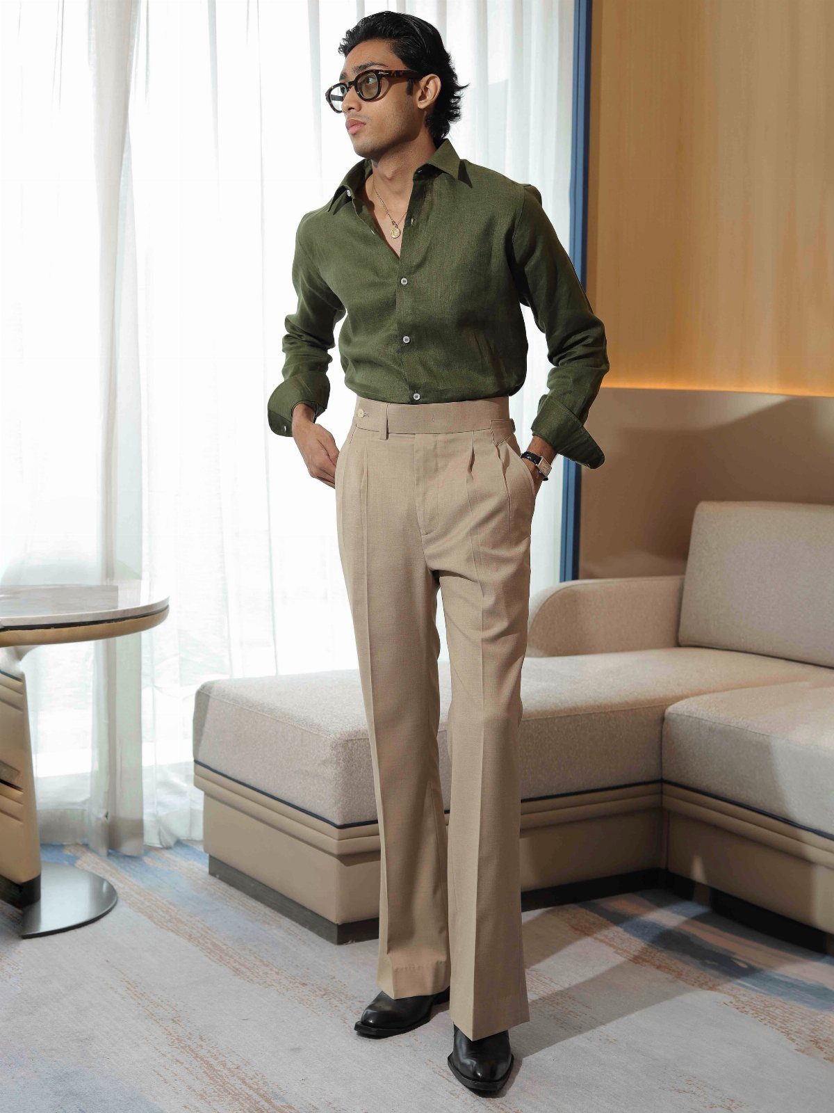 A man wearing a green shirt and beige pleated trousers standing in a bright room.