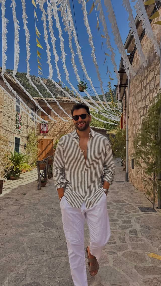 Man in a striped shirt and white trousers walking through a Mediterranean stone village street.