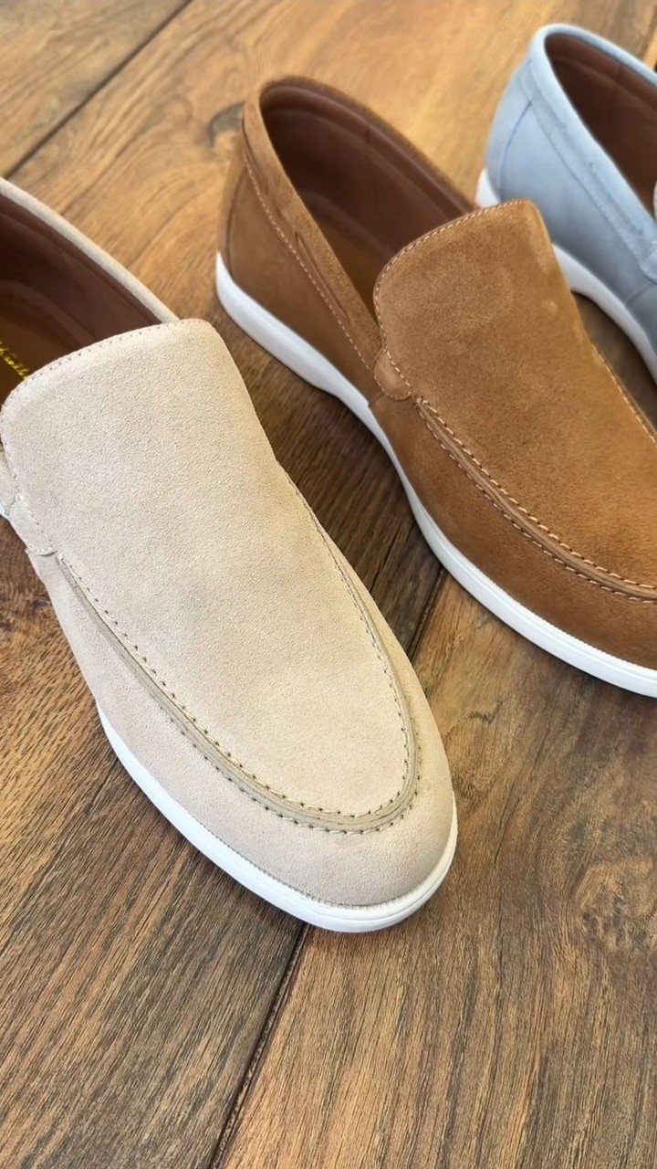 Three suede slip-on loafers in beige, tan, and light blue resting on a wooden floor.