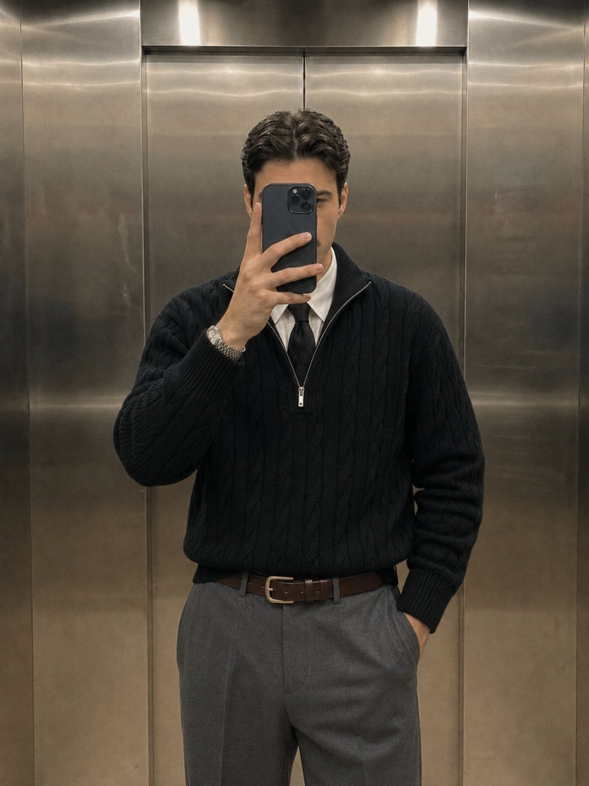 Man in a navy cable-knit quarter-zip sweater, white shirt, tie, and grey trousers taking a selfie.