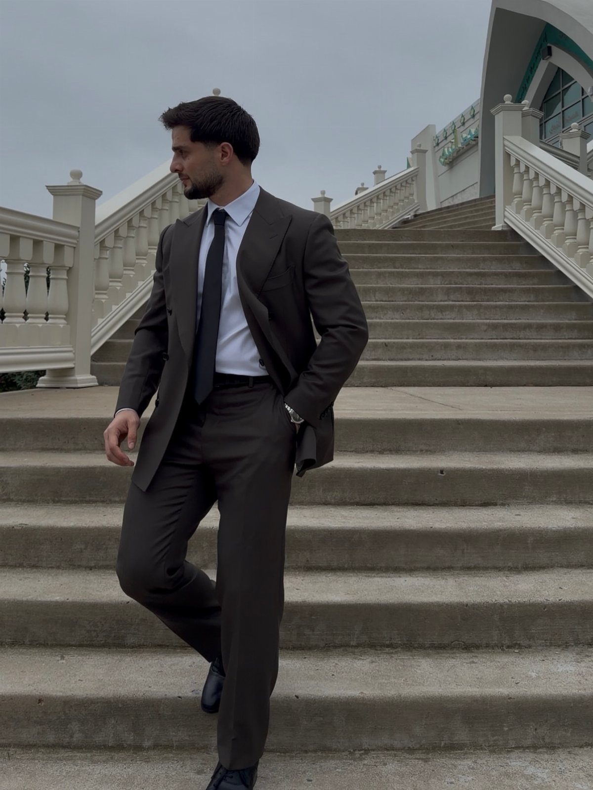 Man in a dark olive green suit posing on stone stairs with white railings.