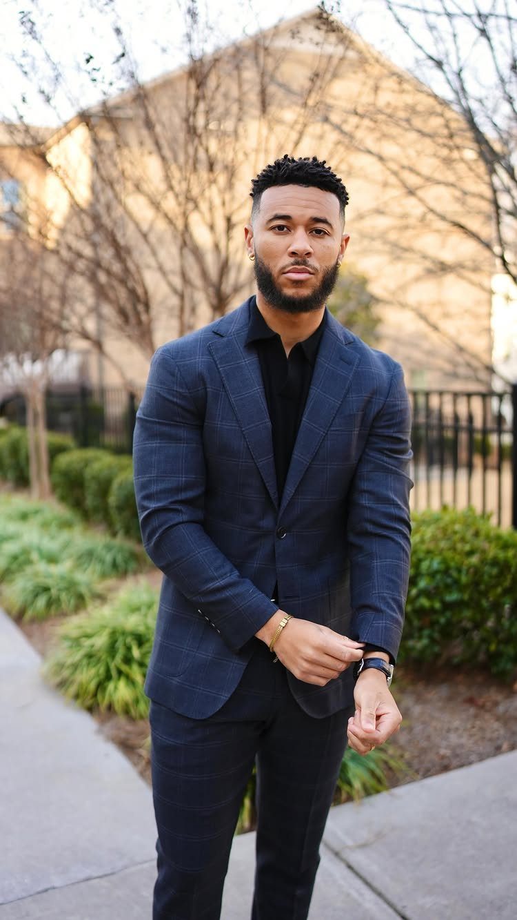 A man wearing a navy blue plaid blazer and black shirt standing outdoors.