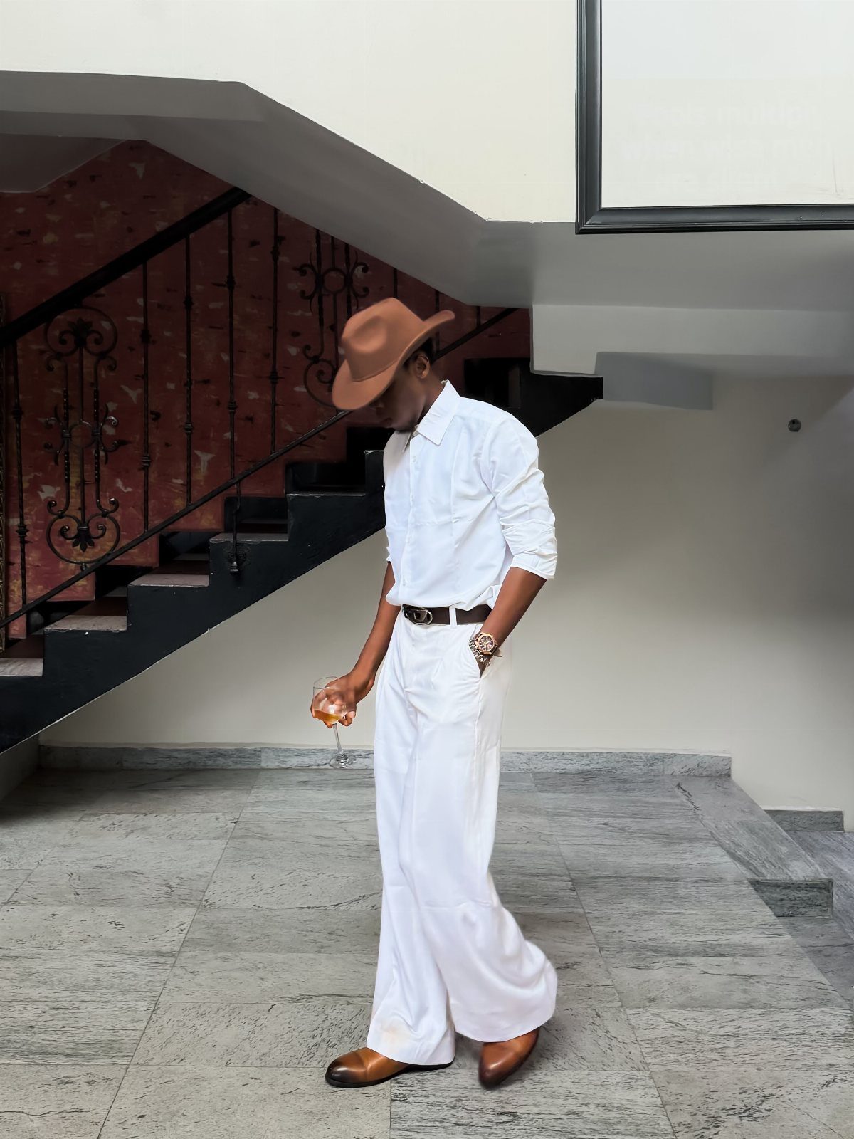 A man in white trousers, white shirt, and brown hat standing near a staircase.