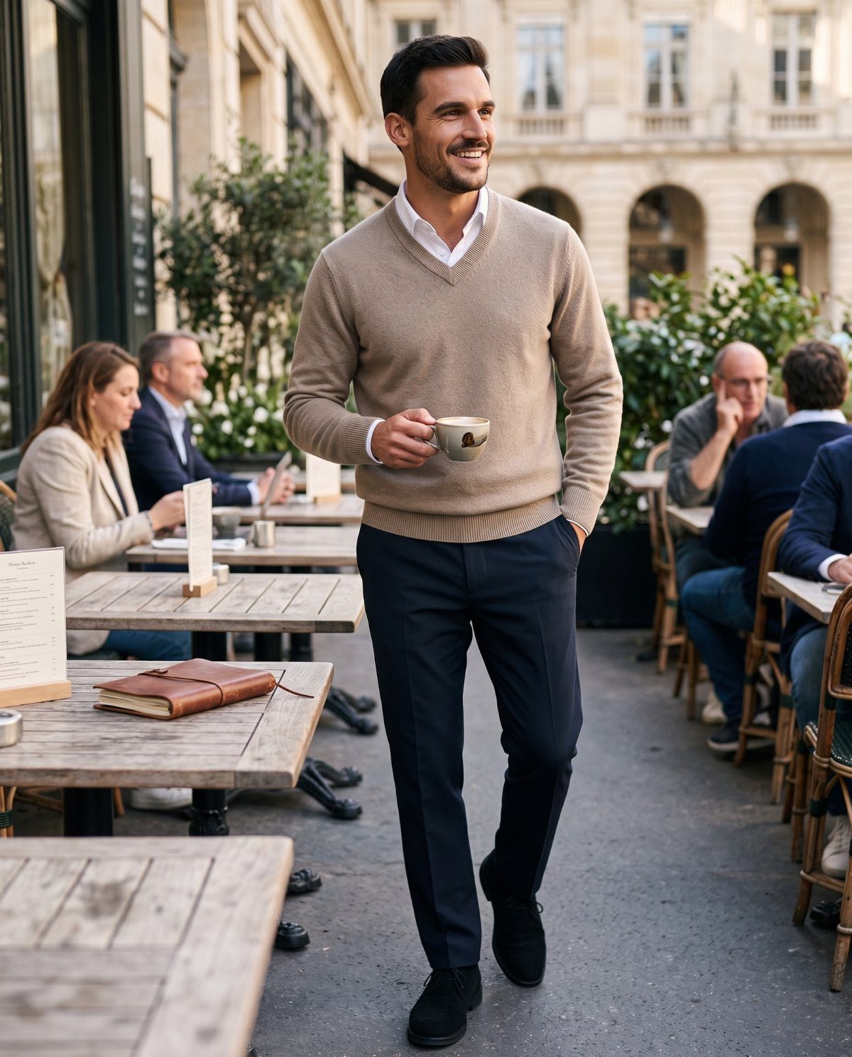 A smiling man in a beige cashmere sweater and navy trousers walking through an outdoor cafe.