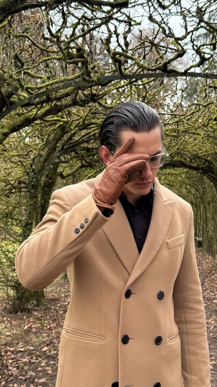 Man in a tan double-breasted coat and brown leather gloves posing among mossy trees.