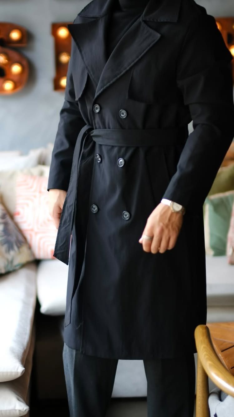 A person wearing a long black double-breasted trench coat with a belted waist.