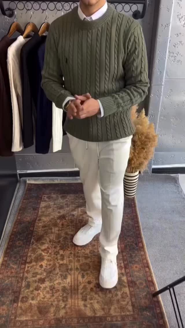 A man wearing an olive green cable knit sweater and white trousers standing on a rug.