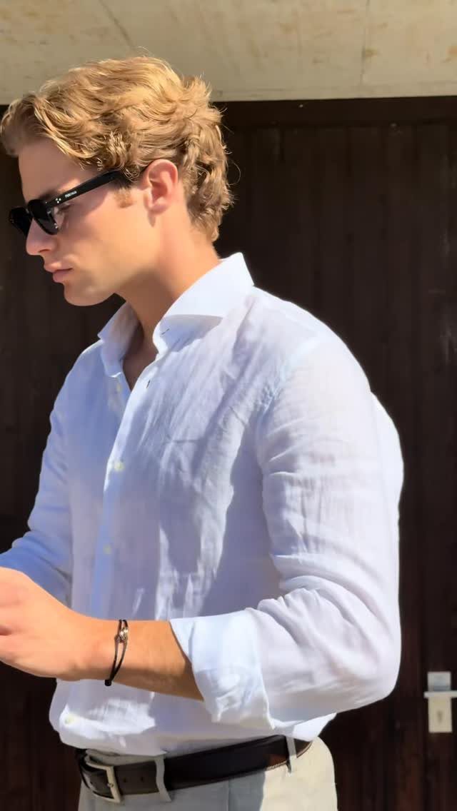 A man wearing a white linen shirt and sunglasses standing against a dark wooden wall.