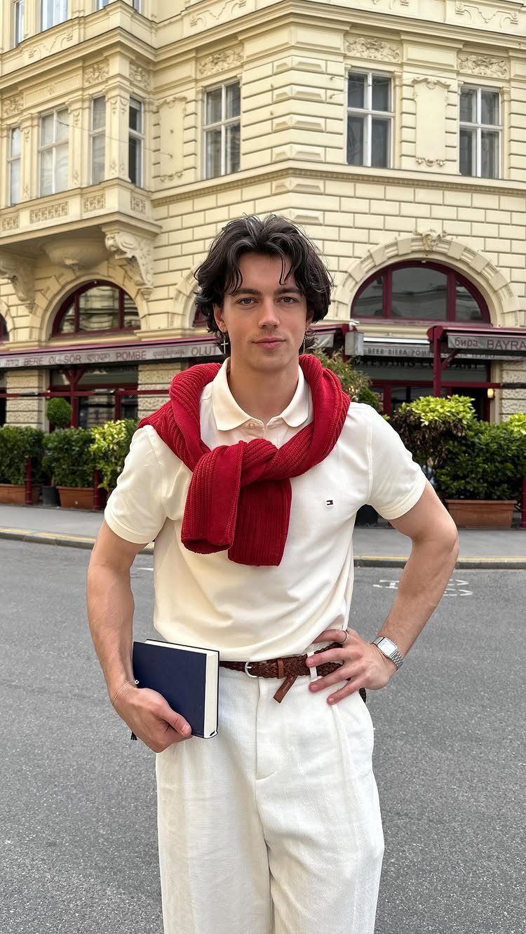 A young man posing in a cream polo, white trousers, and a red sweater draped over his shoulders.