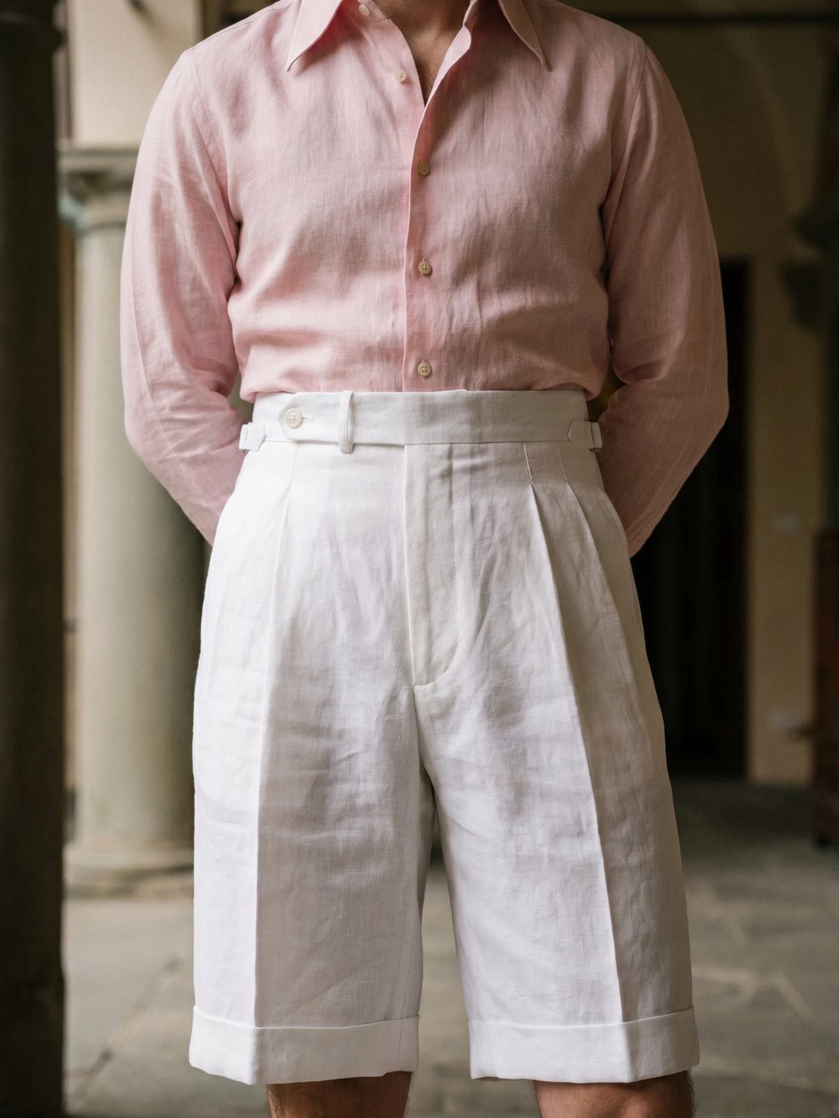 Man wearing a pink linen shirt and white pleated tailored shorts standing outdoors.