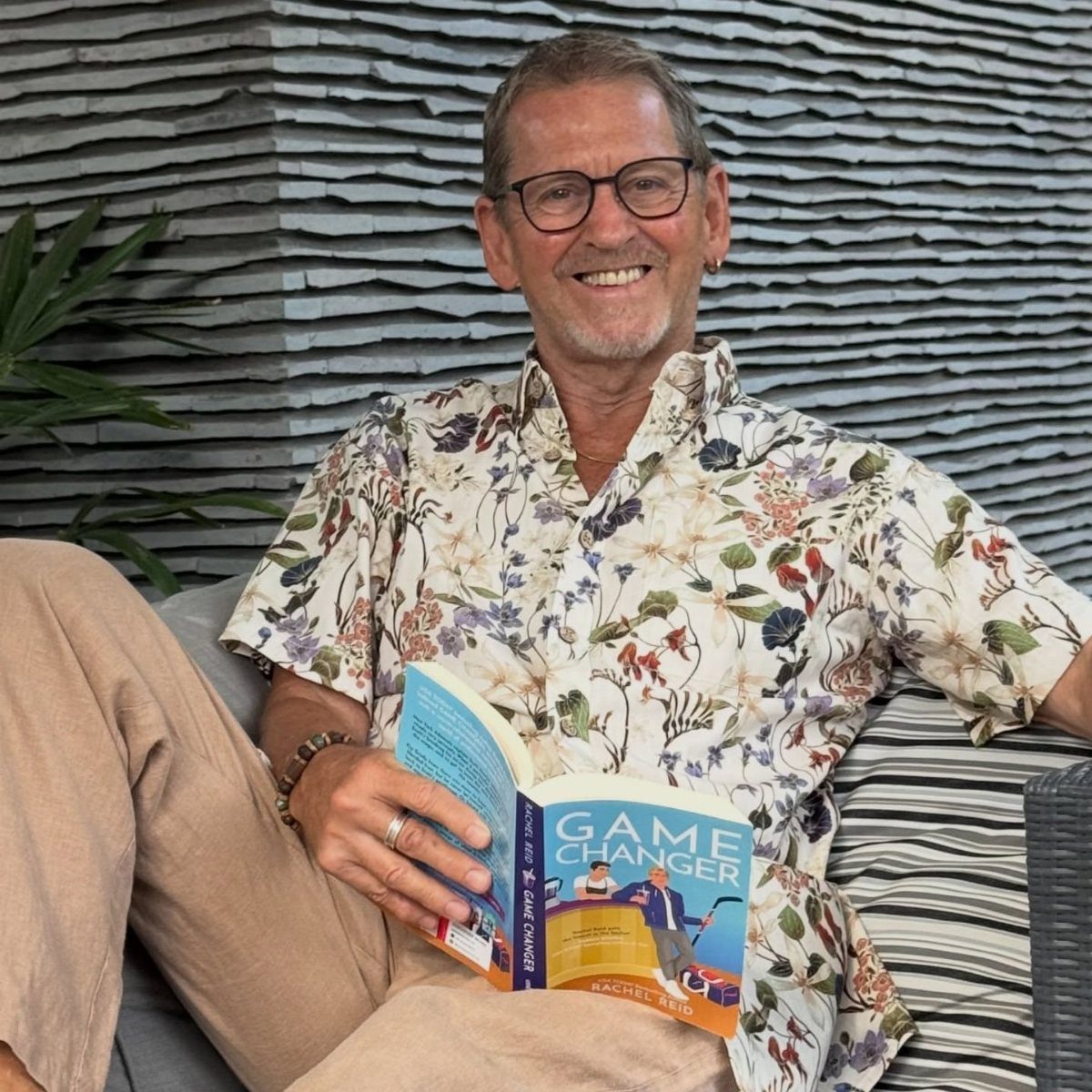 Smiling older man with glasses wearing a floral button-down shirt and holding a book.