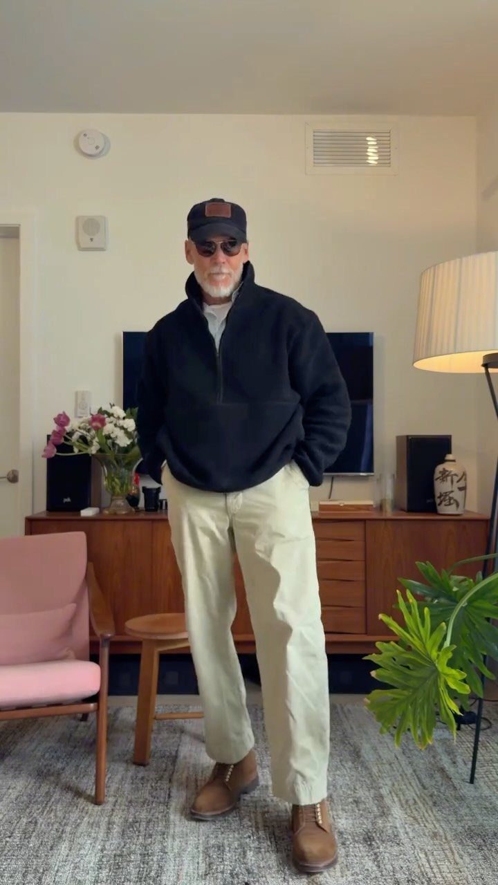 Older man in black fleece, beige pants, and a baseball cap standing in a living room.