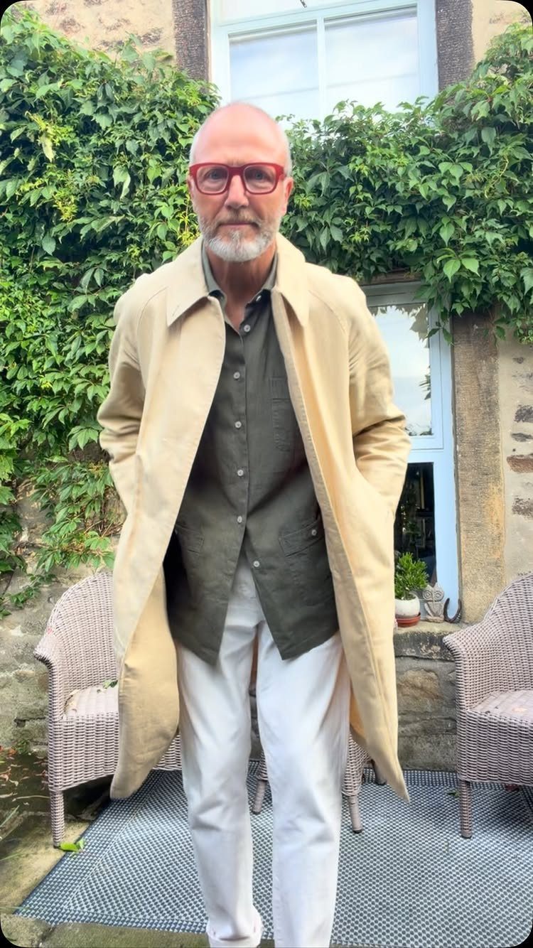 An older man wearing a tan trench coat, olive shirt, and white trousers standing outdoors.