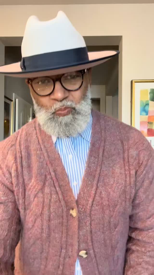 An older man with a grey beard wearing a fedora, glasses, and a knit cardigan.