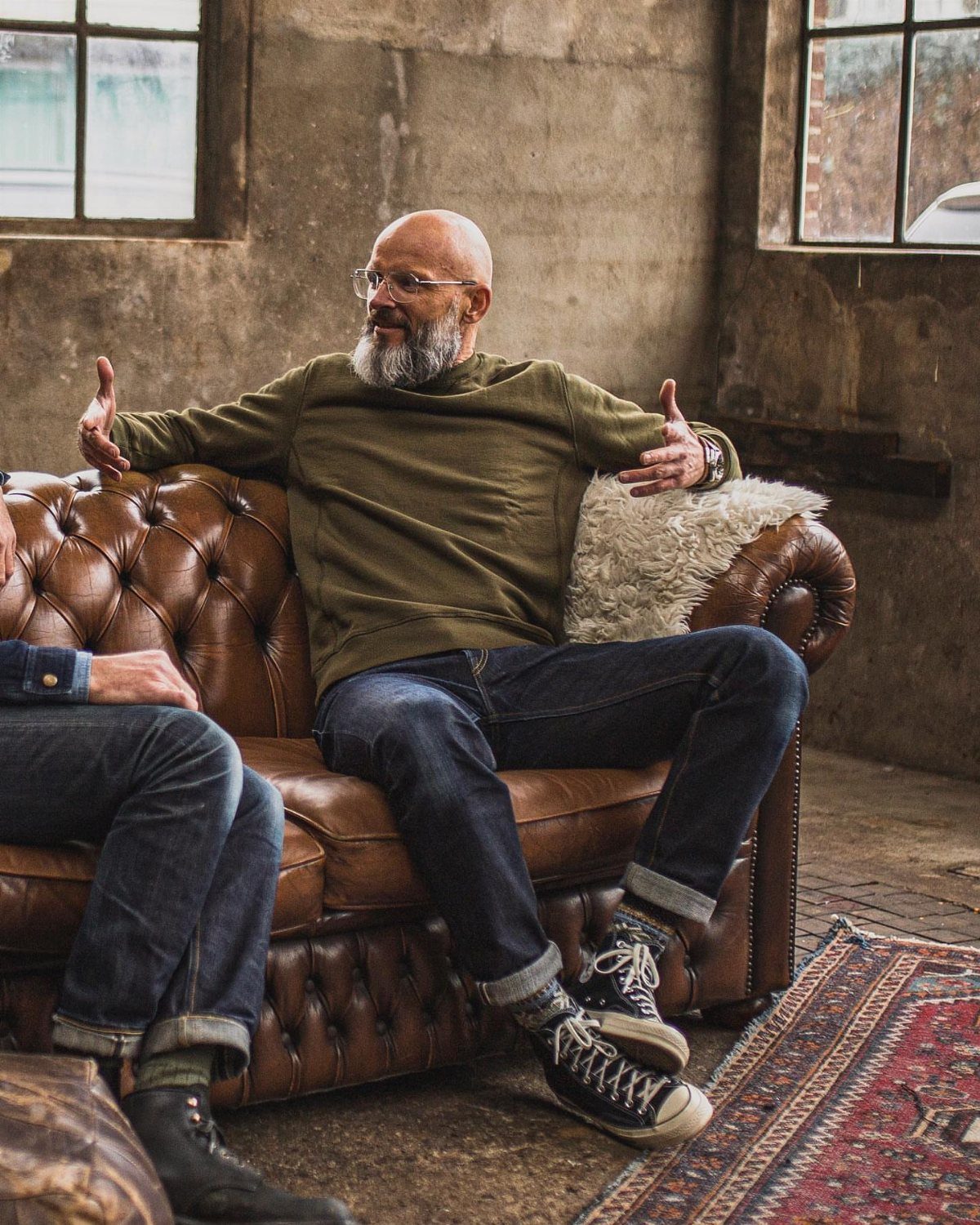 Older man with a grey beard wearing olive green shirt and dark denim jeans sitting on leather sofa.