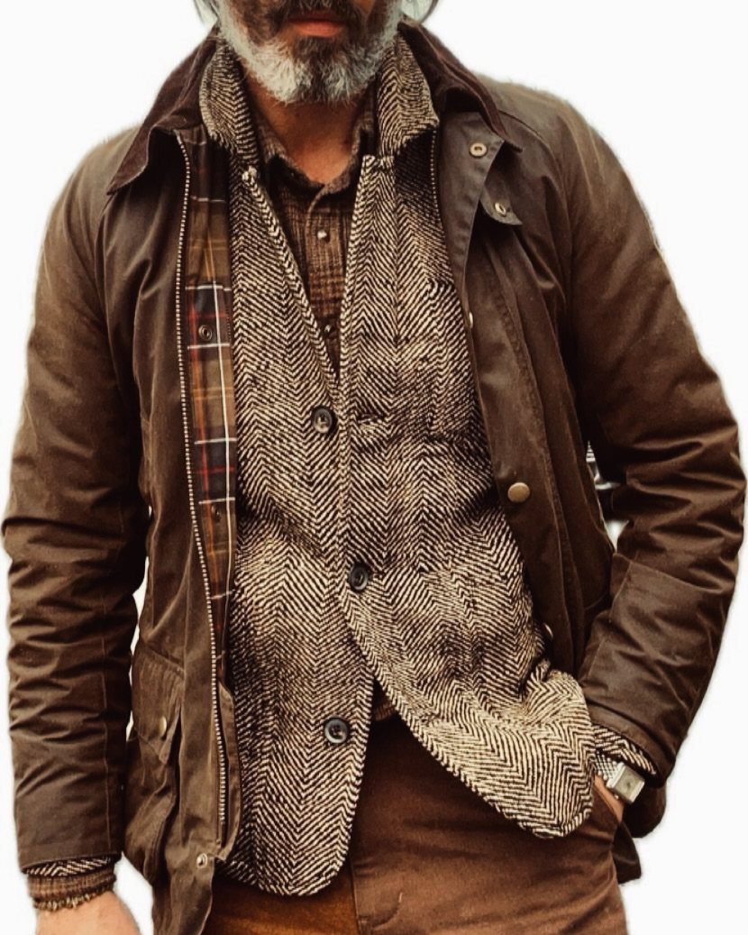 Man wearing a plaid shirt, herringbone waistcoat, and brown waxed jacket for layered styling.