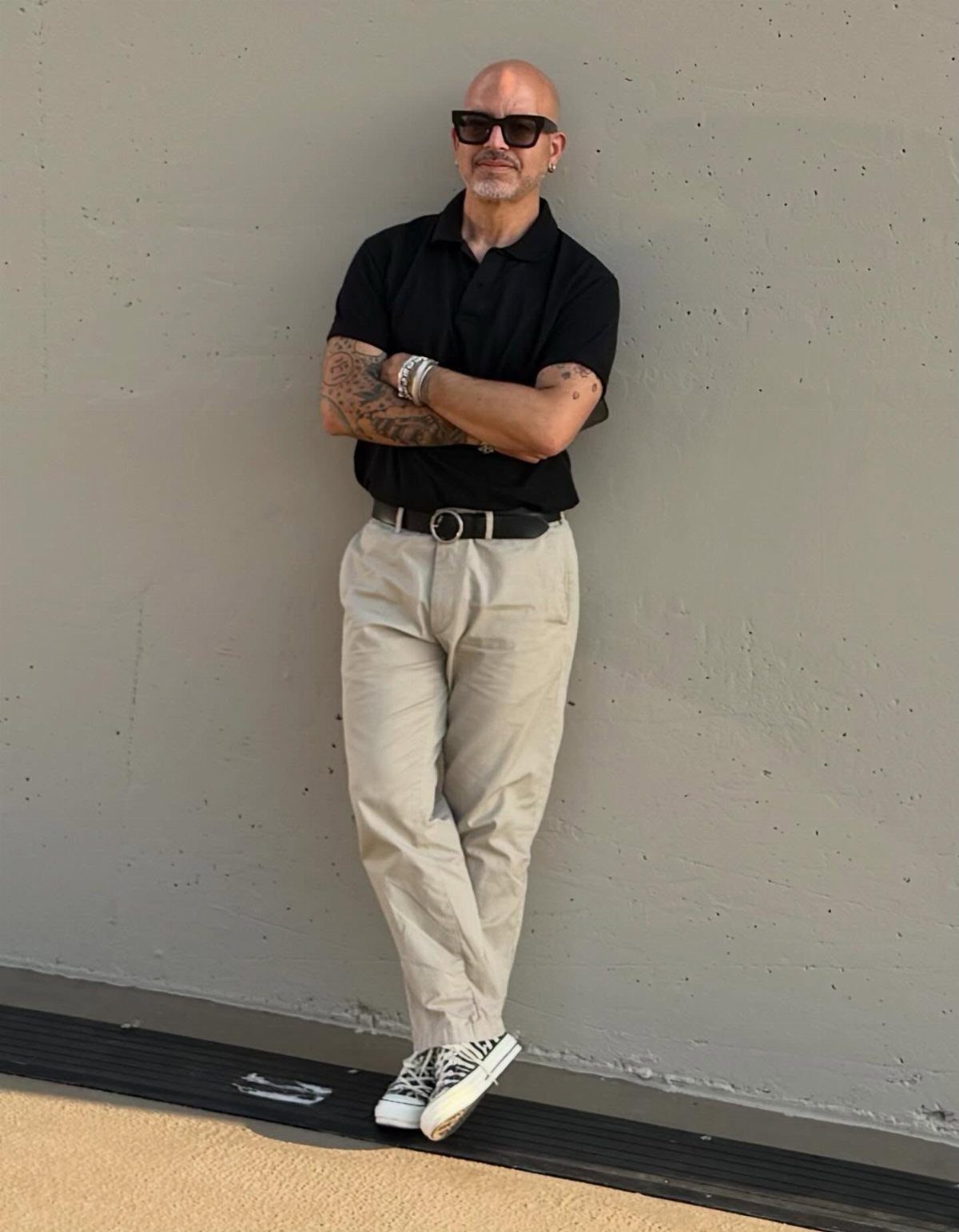 A man in a black polo shirt and beige trousers posing against a grey wall.