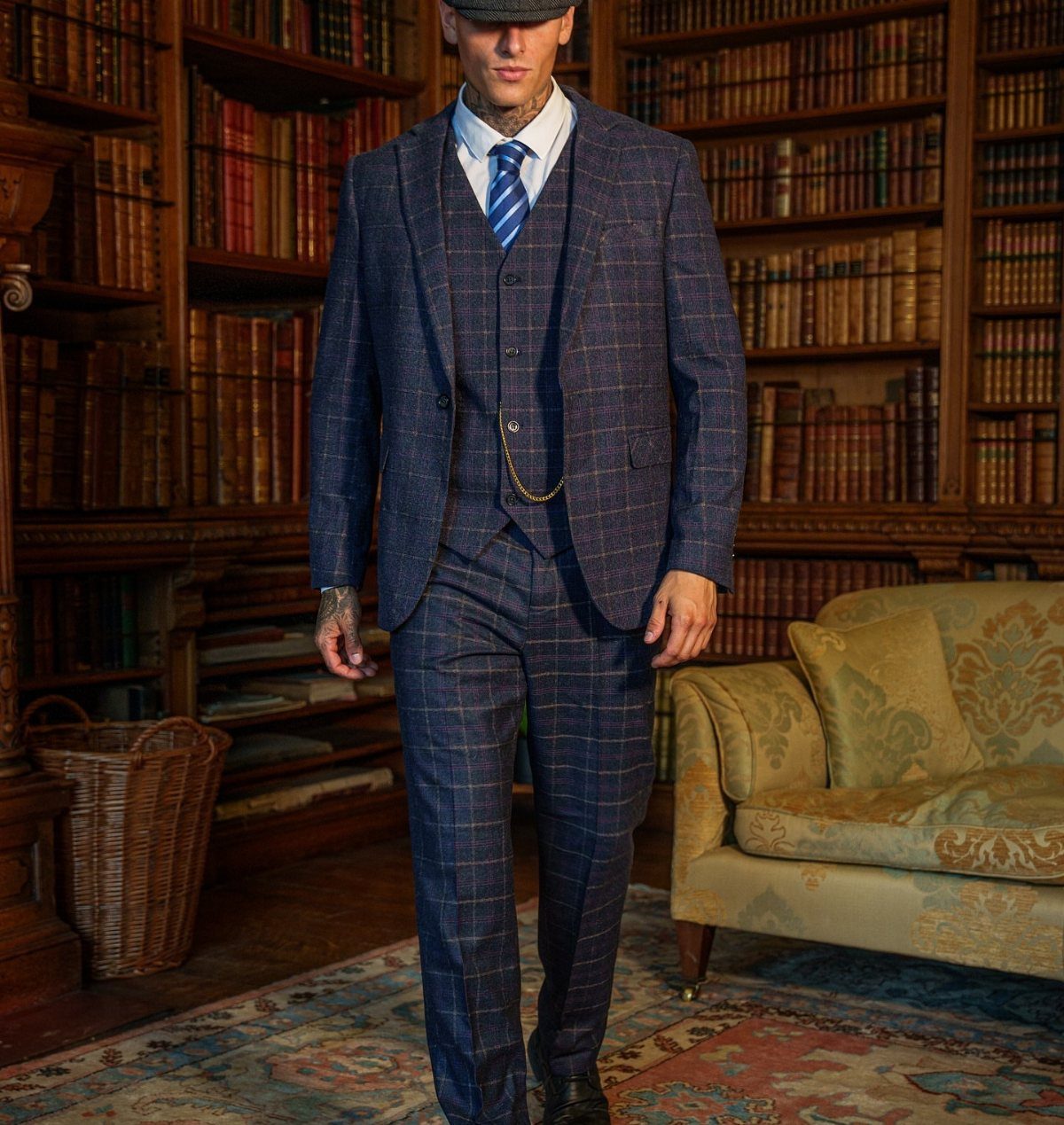 Man wearing a blue plaid three-piece tweed suit and newsboy cap in a library.