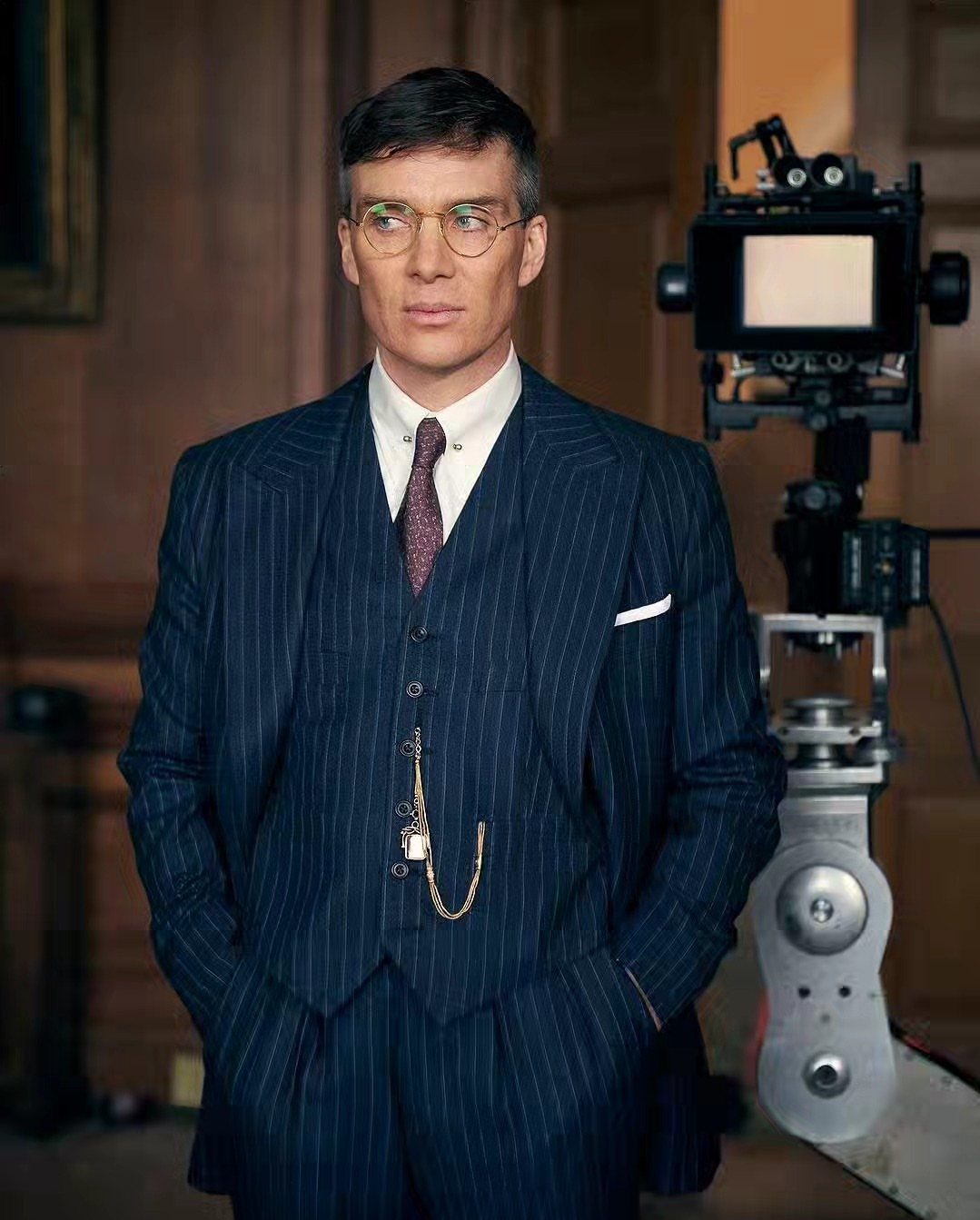 Man in a navy pinstripe three-piece suit with a gold pocket watch chain and round glasses.
