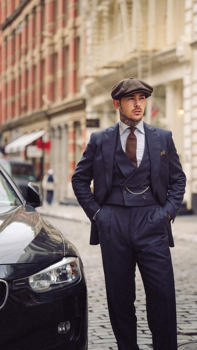 Man in a navy three-piece suit and newsboy cap standing on a city street.