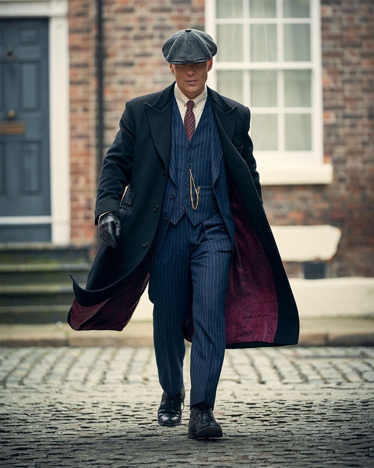 A man in a navy pinstripe three-piece suit and long overcoat walking on cobblestones.