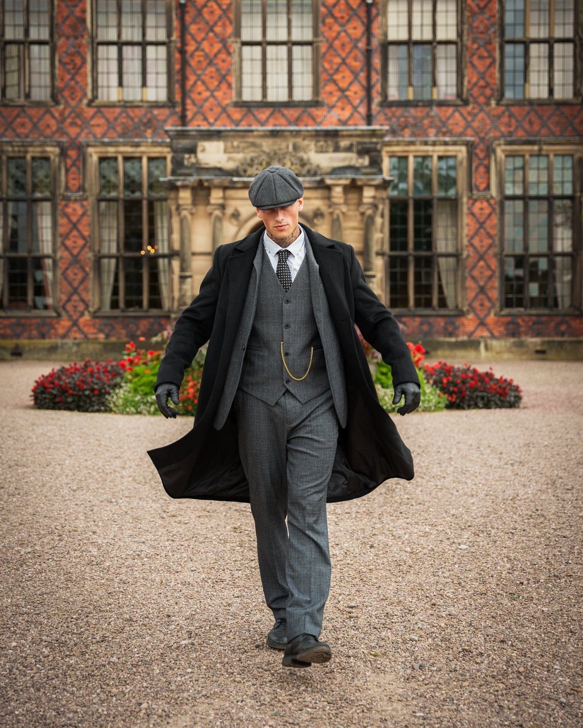 Man in a grey tweed three-piece suit and black overcoat walking outdoors.