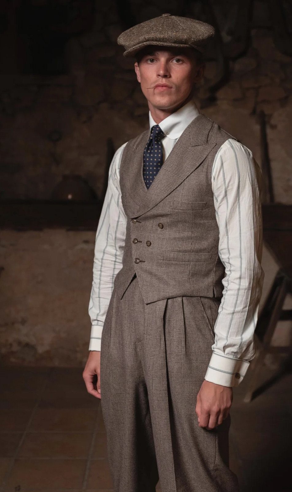 Man in a vintage brown patterned waistcoat, striped shirt, and newsboy cap posing indoors.