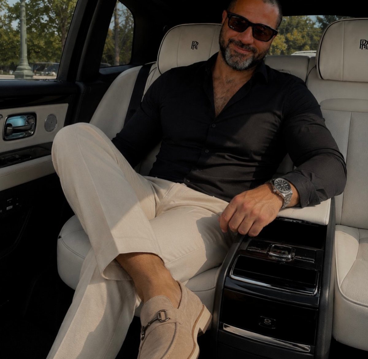 A man in elegant black and beige clothing sitting in a luxury car's leather interior.